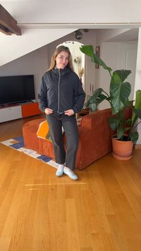 Jauna moteris juodos striukės, tamsių džinsų ir šviesių sportbačių dėvinti, stovi svetainėje su oranžiniu sofa, dideliu kambarinio augalo vazonu ir televizoriumi.