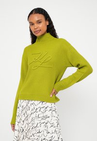 Nő hosszú göndör hajjal, lime zöld bordázott pulóvert visel, melyen kiemelt logó látható, valamint fehér szoknyát fekete írott mintával, pózolva, kezét a csípőjére téve.