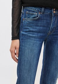 Tummansiniset farkut klassisella istuvuudella, joissa on viisi taskua, kuparinväriset yksityiskohdat ja hienovarainen haalistuma koko kankaassa.