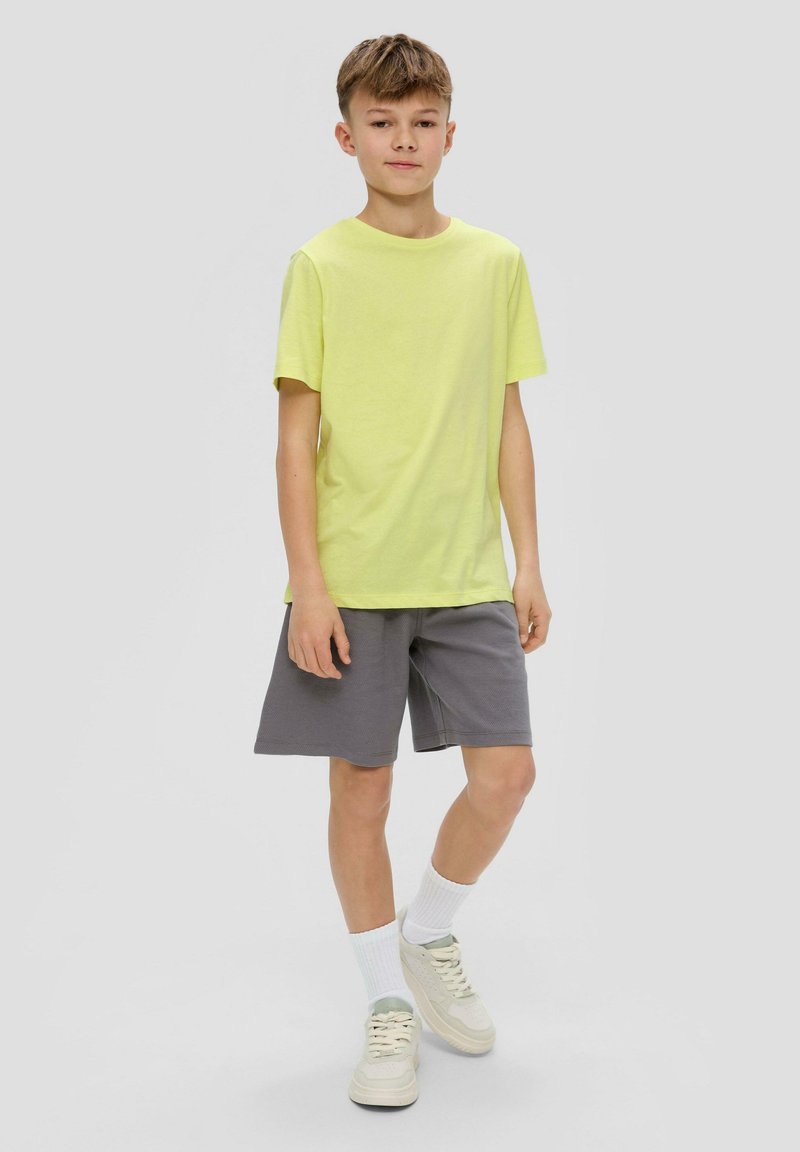 Limetkově zelené bavlněné tričko v kombinaci s šedými šortkami. Oblečení doplňují bílé ponožky a světle zbarvené tenisky.