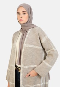 Cardigan en tricot beige avec un motif à carreaux blancs, ouvert sur le devant et doté de poches latérales. Porté sur un haut crème et assorti à un hijab taupe.