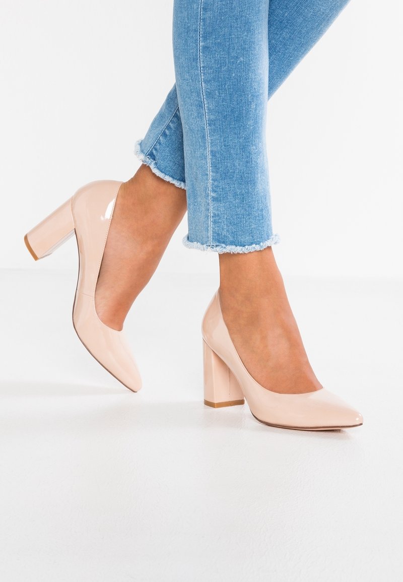 Jambes portant un jean bleu clair à ourlet effiloché et des chaussures à talons carrés en cuir verni nude, sur un sol et un fond blancs.