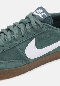 Zelene semišne teniske z belim Nike swooshom, teksturiranimi vezalkami in gumo iz gumija. Povečan pogled poudarja detajle šivanja.