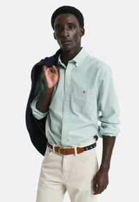 Lichtgroen en wit gestreept overhemd met knopen en een borstzak, gecombineerd met beige broeken en een marineblauwe riem. Een marineblauwe jacket over één schouder gedrapeerd.
