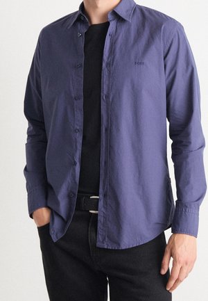 Marineblauwe blouse met knoopsluiting gemaakt van lichte stof, voorzien van een klassieke kraag, slim fit en geborduurd logo op de borst, gecombineerd met zwarte jeans.