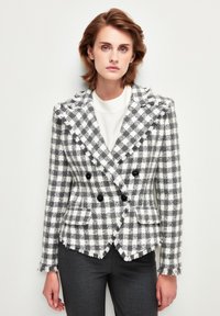 Blazer de doble botonadura en patrón a cuadros en gris y blanco. Tejido texturizado con bordes deshilachados y detalles de botones negros. Corte entallado.
