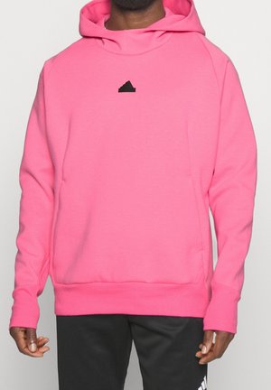 Mann trägt leuchtend pinken Kapuzenpullover mit schwarzem Logo auf der Brust und schwarze Hose vor einfarbigem Hintergrund.