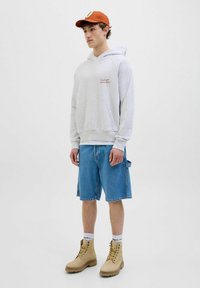 Un sweat à capuche gris clair avec une poche kangourou, associé à un short cargo bleu et des bottes beiges. Accentué par une casquette orange. Fond uni.
