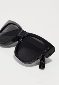 Ochelari de soare negri de mari dimensiuni cu rame din plastic lucios, lentile închise la culoare și mici detalii cu accente argintii pe față.