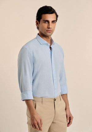 Camisa de manga larga de color azul claro con cuello puntiagudo, frente abotonado y puños remangados, combinada con pantalones beige. Textura de tela suave.