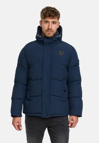 Mężczyzna w granatowej kurtce puchowej z kapturem, stojący na jednolitym jasnym tle, zwrócony przodem z neutralnym wyrazem twarzy.