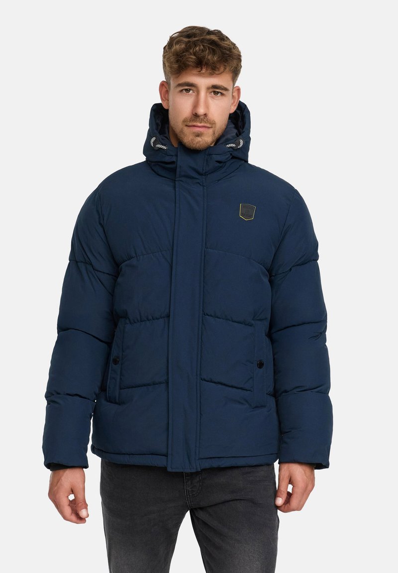 Mężczyzna w granatowej kurtce puchowej z kapturem, stojący na jednolitym jasnym tle, zwrócony przodem z neutralnym wyrazem twarzy.