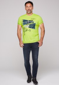 Camiseta verde de manga corta con estampado gráfico que presenta texto en azul. Se lleva con jeans azul oscuro y zapatillas deportivas negras. Diseño sencillo.