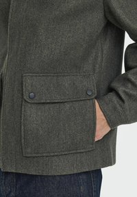 Donkergroene vilten jas met een patchzak met twee zwarte drukknoopjes. Het is gecombineerd met spijkerbroeken, die een gestructureerde afwerking hebben.