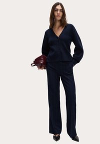 Granatowy kardigan z dekoltem w serek i guzikami z przodu, w zestawieniu z granatowymi spodniami w prążki. Buty mają czubki w kolorze czarnym. Dodatkiem jest burgundowa torba.