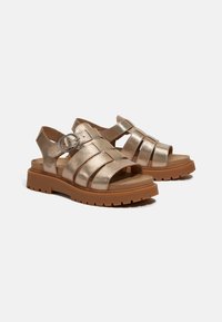 Metallic gold sandals with wide straps, a round buckle, and a thick brown rubber sole. Open back for easy wear. Flat design, textured finish.