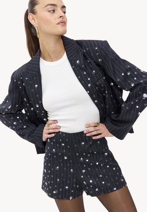 Chaqueta de traje negra con pinstripes y adornos de puntos plateados, combinada con pantalones cortos a juego. Top blanco ajustado por debajo. Textura suave.