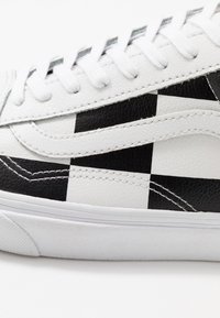 Baskets en cuir blanc avec un motif à damier noir et blanc, bout rond, lacets plats blancs et semelle en caoutchouc texturée.