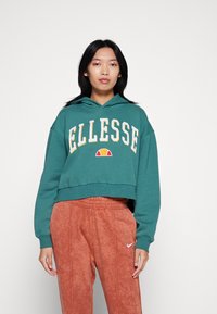 Felpa verde acqua corta con grande logo "ELLESSE" in bianco e giallo, dotata di tasca frontale a marsupio e cappuccio con cordino, abbinata a pantaloni color ruggine.