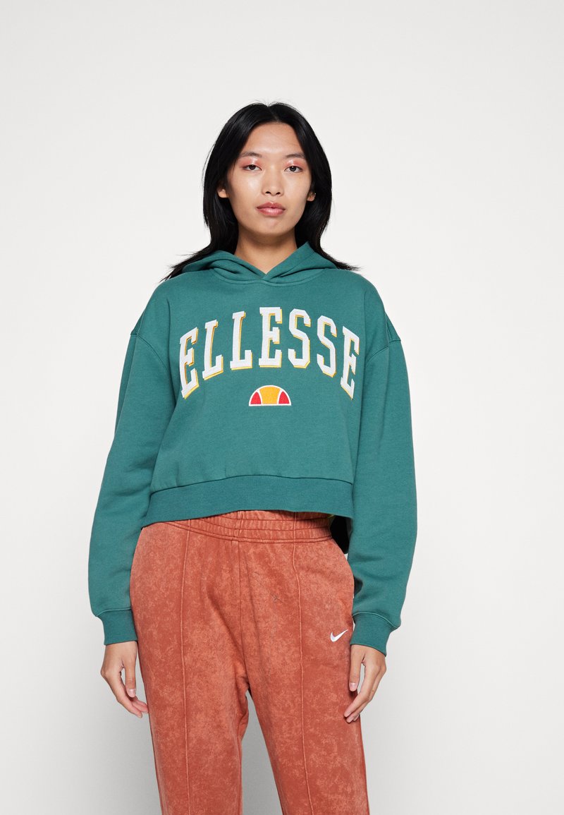 Felpa verde acqua corta con grande logo "ELLESSE" in bianco e giallo, dotata di tasca frontale a marsupio e cappuccio con cordino, abbinata a pantaloni color ruggine.
