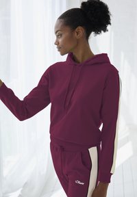 Bordeaux hoodie met verstelbare trekkoord, voorzien van contrasterende witte zijstrepen. Zachte stof, ribgebreide boorden en geborduurd logo op de borst.