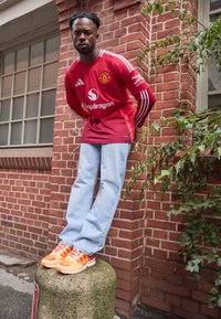 Raudona "Manchester United" marškinėliai su baltais dryžiais, šviesiai mėlyni džinsai ir oranžiniai sportbačiai, stovintis ant betono stulpo prieš plytų sieną.