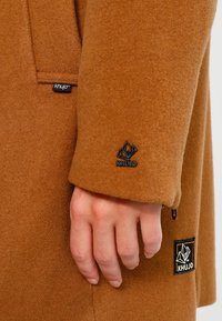 Brauner Wollmantel mit weicher Textur, mit auffälligen KHUJO-Labels am unteren Ärmel und an der Seitentasche. Hand, die auf dem Stoff ruht.