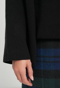 Black knitted sweater with wide sleeves, paired with a blue and green plaid skirt. Soft texture visible on fabric.