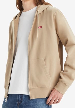 Haut à capuche beige zippé en tissu doux, avec une poche avant, des cordons de serrage et un petit logo rouge Levi's sur la poitrine gauche.