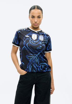 Femeie cu păr dat pe spate purtând un tricou de fotbal al Argentinei cu model negru și albastru și pantaloni negri, stând cu o mână în buzunar.