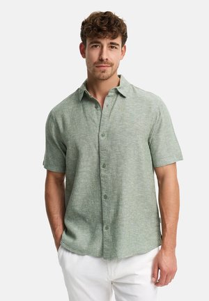 Młody mężczyzna o kręconych włosach, ubrany w jasnozieloną koszulę na krótki rękaw z guzikami i białe spodnie, stojący z rękami w kieszeniach.