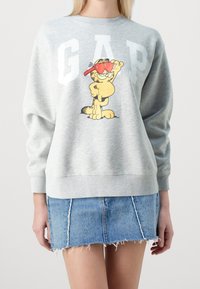Sudadera gris con grandes letras blancas "GAP" y un diseño de un gato de dibujos animados. La tela es suave, con un corte relajado y puños acanalados. Falda de mezclilla debajo.