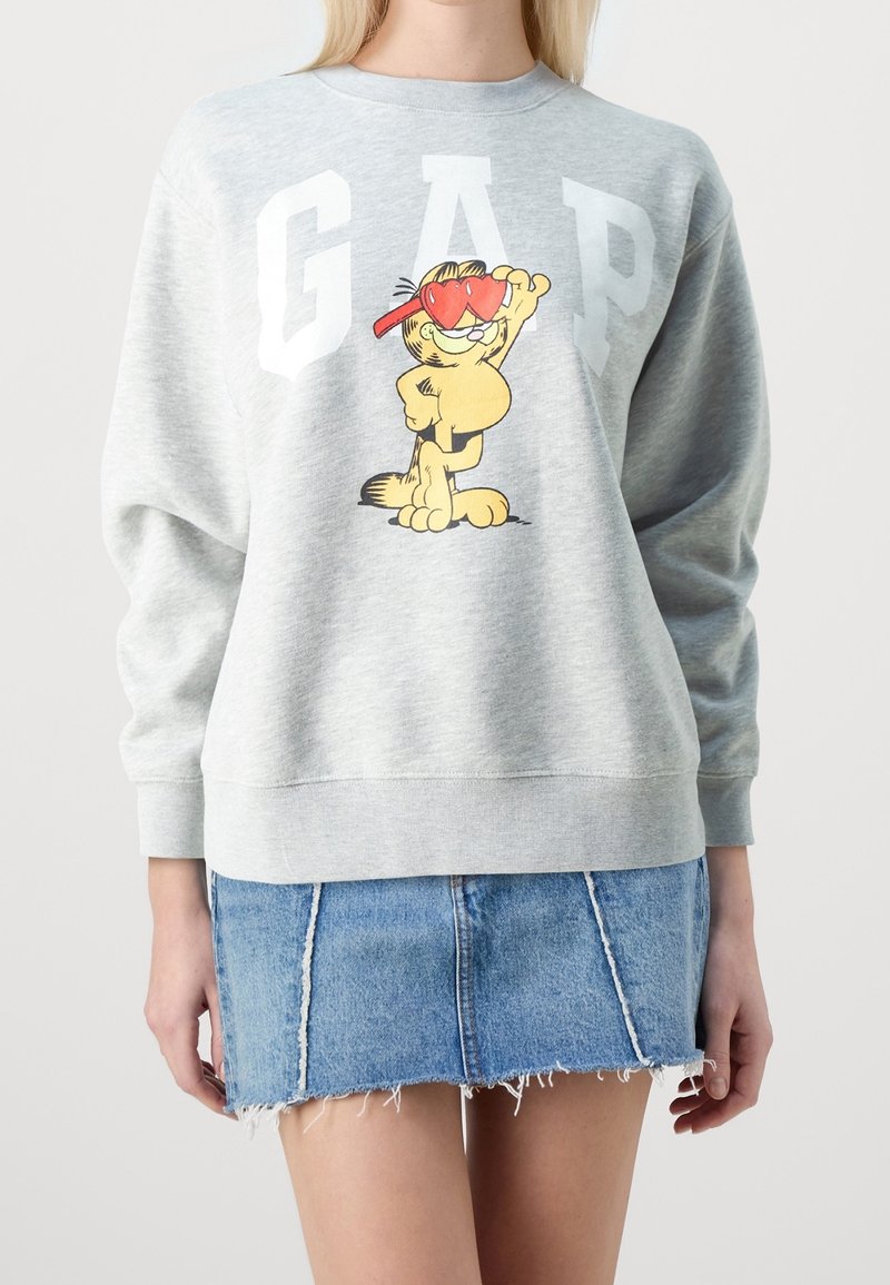 Sudadera gris con grandes letras blancas "GAP" y un diseño de un gato de dibujos animados. La tela es suave, con un corte relajado y puños acanalados. Falda de mezclilla debajo.