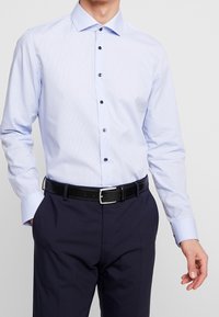 Vaaleansininen pitkähihaista nappipaitaa, jossa on ruutukuvio, mustat napit ja levitetty kaulus, yhdistettynä tummiin housuihin ja mustaan vyöhön.