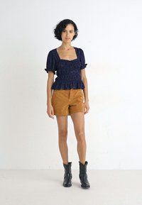 Marineblauw peplum top met korte pofmouwen en geplooide textuur, gecombineerd met bruine suede shorts en zwarte enkelboots.