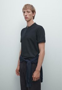 Sort kortærmet henley skjorte med en struktureret ribbet stof; parret med mørke bukser og en marineblå sweater bundet om livet.