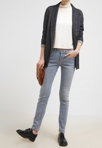 Šedý kardigan přes bílý top, doplněný světle modrými skinny džíny a černými šněrovacími botami. Drží hnědou kabelku.