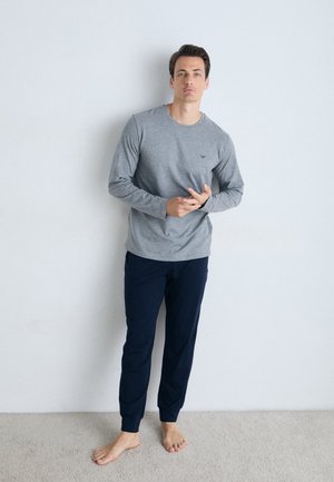 Gray long-sleeve shirt and navy pants, made of soft fabric; shirt features a small logo on the chest. Model stands barefoot on carpet.