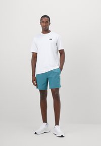 Mężczyzna stojący, ubrany w białą koszulkę Adidas, turkusowe szorty i białe buty Adidas na tle jasnego, jednolitego tła.