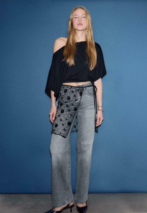 Čierny top s odhalenými ramenami, svetlošedé rifle s čiernou kvetinovou čipkou na jednej strane a čierne lodičky, na modrom pozadí.