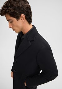 Cappotto di lana nera con finitura testurizzata, design a singolo petto, rever a punta e due tasche frontali. Il modello è visto di profilo.
