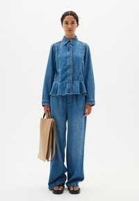 Blauwe denim peplumjacket met lange mouwen en kraag, gecombineerd met een broek met wijde pijpen. Model houdt een tan tote bag vast; weergegeven tegen een witte achtergrond.