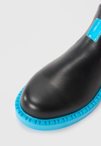 Black leather shoe with a rounded toe and contrasting blue rubber sole featuring textured ridges and a blue accent label on the side.