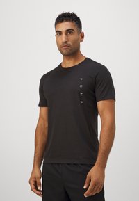 Camiseta preta de manga curta feita de algodão, com um corte justo e um logótipo branco vertical do lado esquerdo. Textura suave.