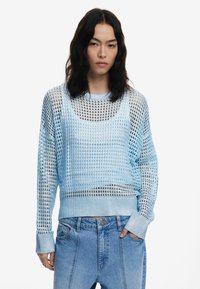 Hellblauer Mesh-Pullover mit lockerer Passform und weiten Ärmeln. Mit gerippten Bündchen und einem strukturierten Muster. Kombiniert mit hochgeschnittenen Jeans.