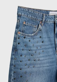 Jean en denim bleu avec de petits clous métalliques décorant la poche avant et une étiquette Stradivarius visible sur la ceinture.