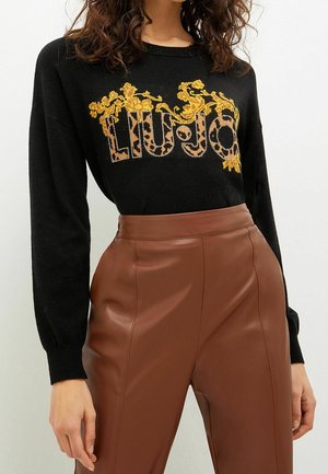 Woman wearing black sweater with "LIU JO" in leopard print and yellow floral embroidery, paired with high-waisted brown leather pants.