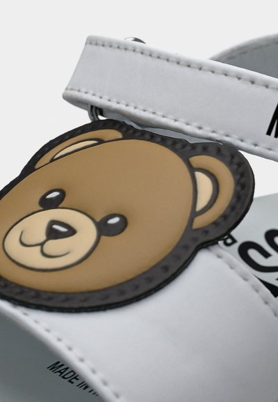 Λευκό σανδάλι παιδικό με καφέ καρτούν αρκουδάκι στο λουράκι, με ορατή ραφή και τυπωμένο κείμενο στο εσωτερικό.