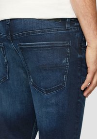 Jeans en denim bleu foncé avec une texture lisse, présentant un design de poche arrière et de légers détails usés le long des bords.