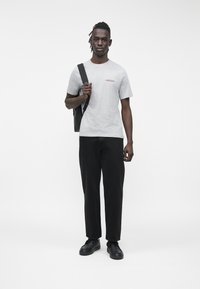 Szürke pamut póló piros felirattal, fekete bő szárú nadrág és fekete tornacipő. A modell fekete táskát visz, fehér háttér előtt áll.